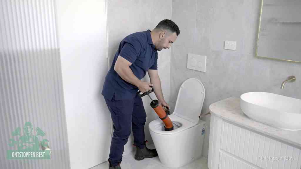 Een monteur buigt over een wandcloset en duwt met de hand een persluchtontstopper in de toiletpot om de verstopping handmatig los te maken in Best