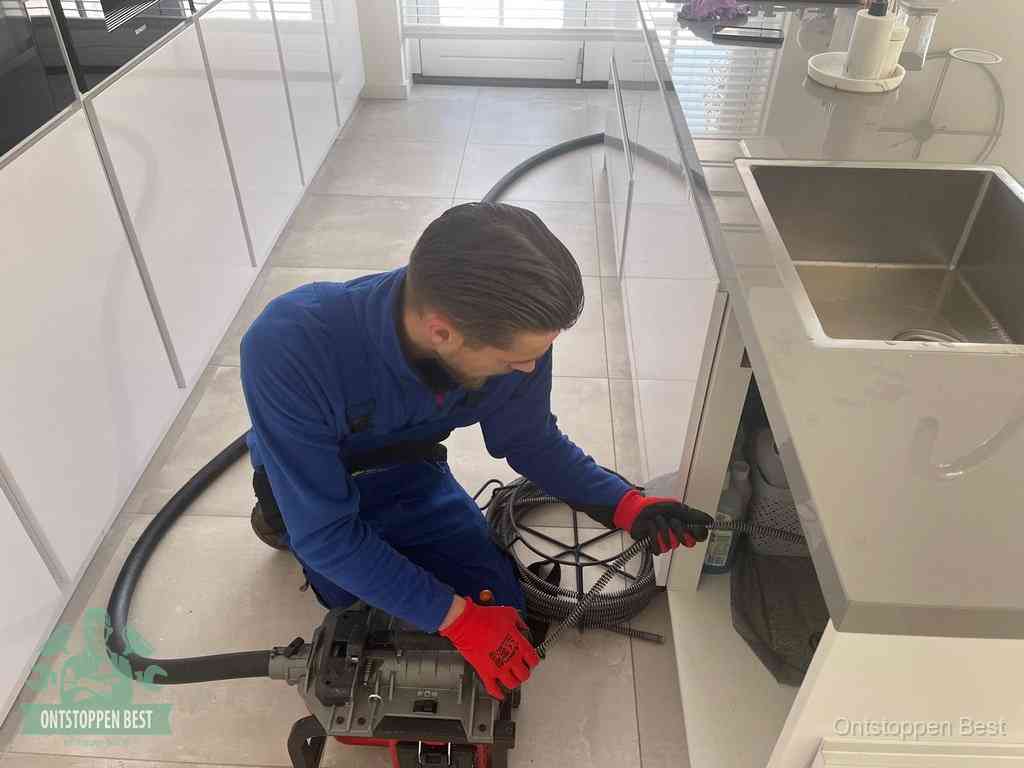 Een monteur ontstopt de keukenafvoer onder de spoelbak met een elektrische veermachine in Best