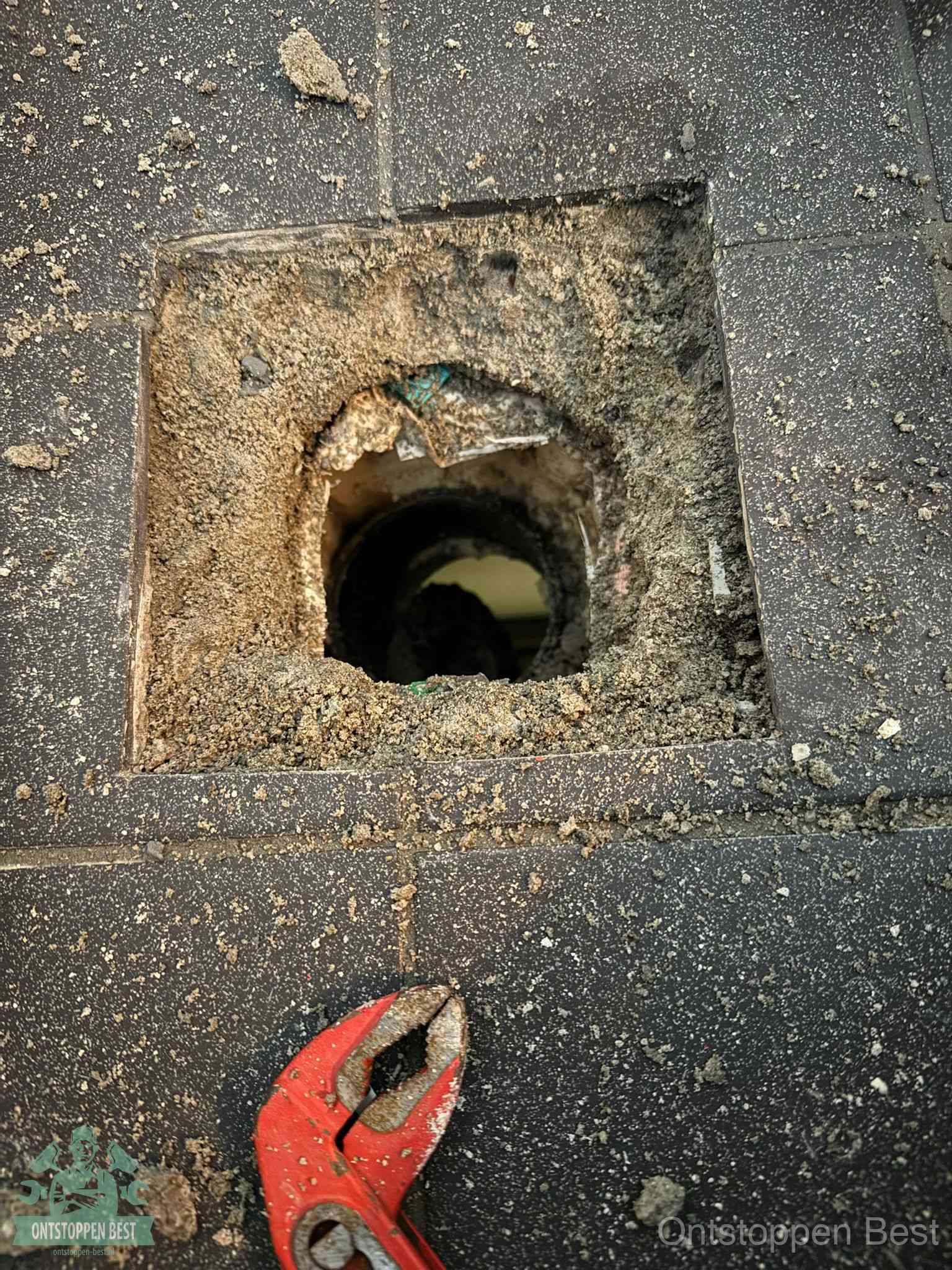 Een geopend rioolputje in een betontegelvloer met losgemaakt rooster en een zichtbare ronde afvoerbuis, het achterhalen waar de afvoerverstopping zit in Best