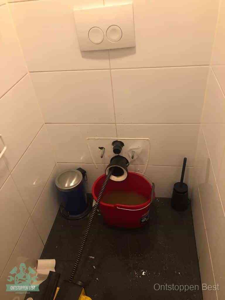 Een ontstopte wand-wc met verwijderde toiletpot waar een ontstoppingsveer in de wanduitgang verdwijnt, het achterhalen waar de verstopping zit in Best