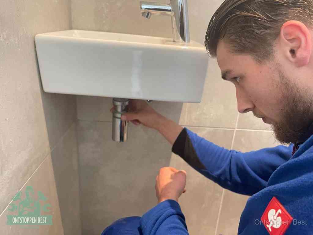 Een monteur draait met de hand de zwanenhalssifon onder een fonteinwasbak in de toiletruimte los om de bron van rioollucht in Best te vinden