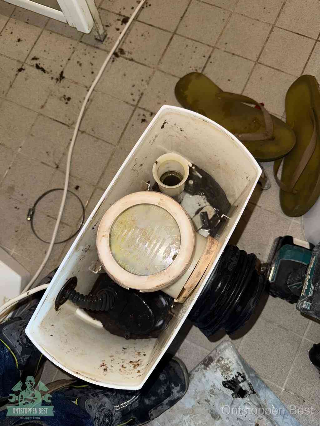 Een gedemonteerde toiletpot op de vloer met ontstoppingsveer en gereedschap ernaast, het verwijderen van een vastzittend voorwerp uit de wc in Best