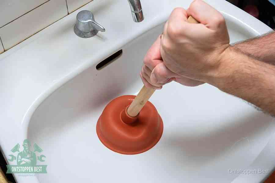Twee handen gebruiken een rode rubberen plopper op een sanitair-afvoer om het toilet te ontstoppen in Best