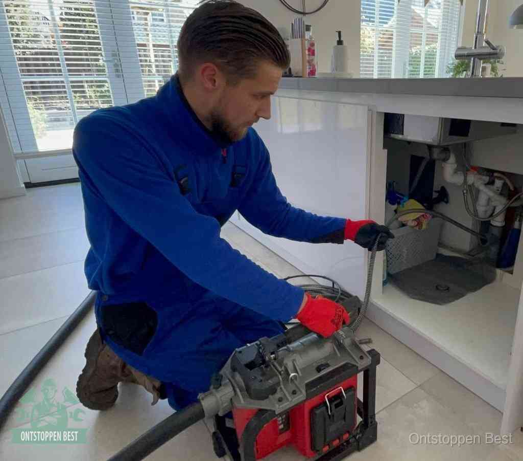 Een monteur met rode handschoenen knielt in een keuken en voert een elektrische ontstoppingsveer in tijdens het professioneel afvoer ontstoppen in Best