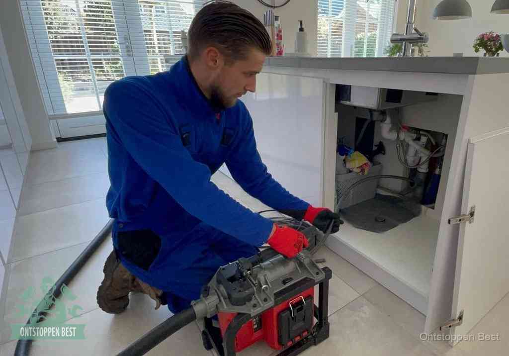 Een monteur voert een professionele elektrische ontstoppingsmachine in de afvoer onder de spoelbak tijdens het gootsteen ontstoppen in Best