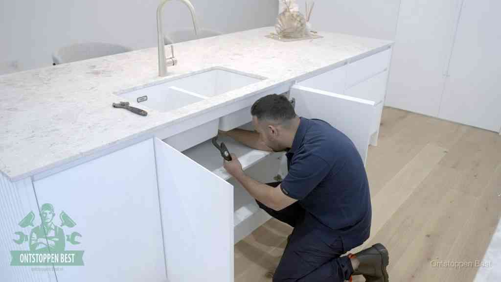 Een man knielt voor een open keukenkast onder de spoelbak om zelf de verstopte afvoer aan te pakken in Best